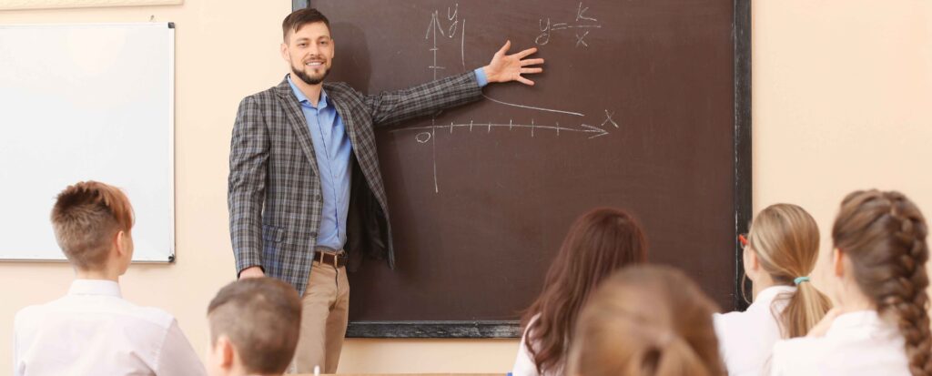 male teacher standing at front of class pointing to the chalkboard while teaching class
