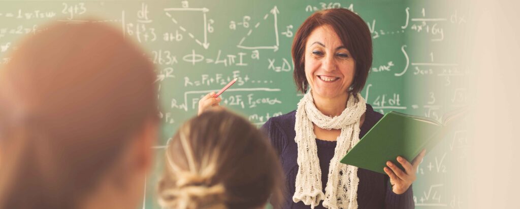 female teacher standing at front of class in front of green chalkboard talking to the class. 