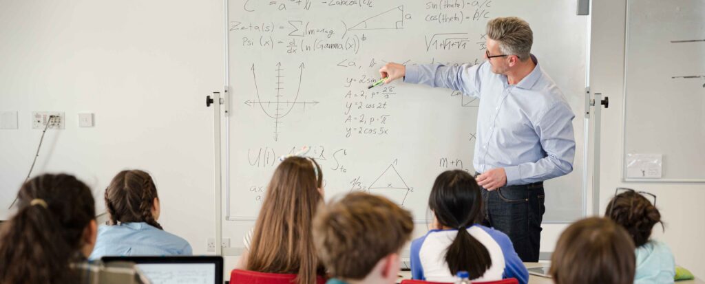 male math teacher standing at the front of class pointing to an equation on the board. 