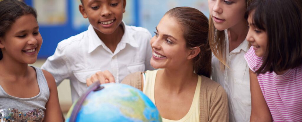 teacher sitting a table surround by her student as she points to a section on a globe. 