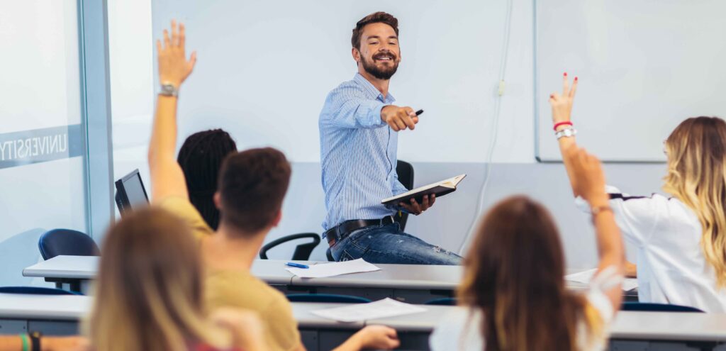 teacher leaning on a desk at the front of class pointing to a student who with their hand raised