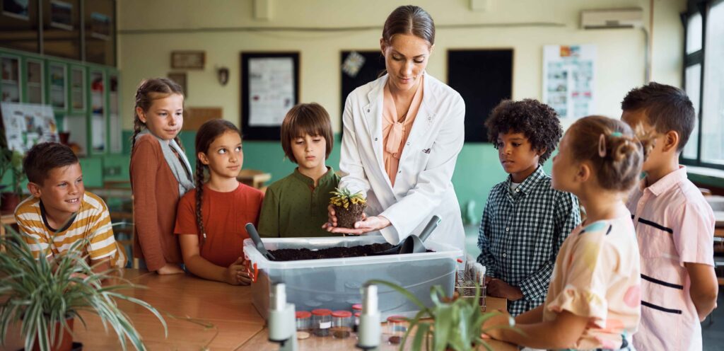 teacher in a lab coat holding a plant as her students gather around her