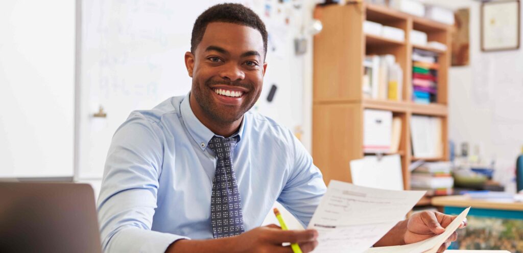 teacher sitting at his desk with a piece of paper in his hands smiling for a picture